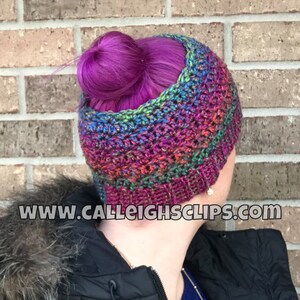 May include: A colorful crochet hat with a bun hole. The hat is made with a variety of colors, including purple, blue, green, and red. The hat is worn by a person with purple hair. The website www.calleightsclips.com is visible in the image.