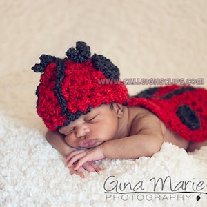 May include: A newborn baby wearing a red and black ladybug crochet costume. The baby is sleeping on a white fluffy blanket. The costume has black spots and a black head with antennae.