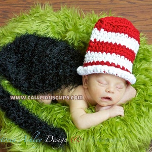 May include: A newborn baby wearing a red and white striped crocheted hat, sleeping on a green fluffy surface. The baby is wearing a black fluffy tail.