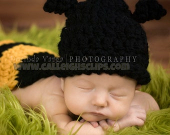 May include: A newborn baby wearing a black crocheted bee hat with yellow and black striped pants, sleeping on a green fuzzy surface.