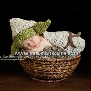 Puede incluir: Un bebé recién nacido con un gorro y una manta de crochet Yoda verde y marrón, durmiendo en una cesta de mimbre marrón.