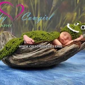 May include: A newborn baby wearing a green crocheted frog hat and a green crocheted frog costume is sleeping on a piece of driftwood. The baby is lying on its side with its head resting on the wood. The background is a blurry green and brown.
