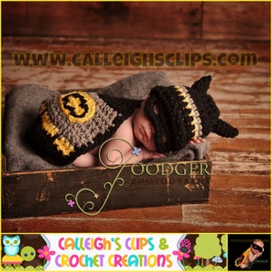 Puede incluir: Un bebé recién nacido con un disfraz de Batman de crochet negro y amarillo, durmiendo en una caja de madera. El disfraz incluye una capa y una máscara. El bebé está acostado de lado con la cabeza apoyada en el brazo. La caja es de madera oscura y tiene un acabado desgastado. El fondo es un suelo de madera.