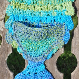May include: A crocheted mermaid tail blanket in shades of blue and green. The tail is folded in half, showing the inside of the blanket. The blanket is hanging on a wooden bench.