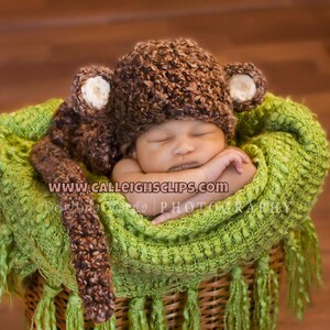 May include: A newborn baby wearing a brown crocheted monkey hat with large ears, sleeping in a green crocheted basket with fringe.