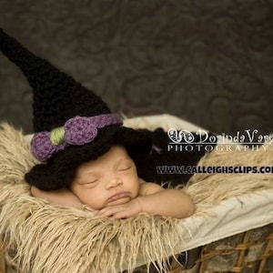 May include: A newborn baby wearing a black crocheted witch hat with a purple bow and green accents, sleeping in a wicker basket lined with faux fur.