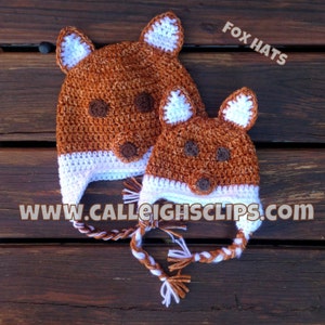 May include: Two crocheted fox hats. The hats are brown with white accents on the ears and around the face. The hats have brown eyes and noses. The text "FOX HATS" and "WWW.CALLEIGHCLIPS.COM" are visible.