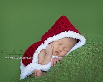 May include: A newborn baby wearing a red and white crocheted Santa hat and matching outfit is sleeping on a green textured surface.