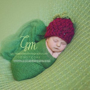 May include: A newborn baby wearing a green swaddle and a red crocheted strawberry hat is sleeping on a green knitted blanket. The baby's eyes are closed and the baby's mouth is slightly open. The baby's hands are tucked under the chin.