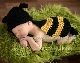 May include: A newborn baby wearing a black and yellow crocheted bee costume with a black crocheted bee hat. The baby is lying on a green fuzzy surface.