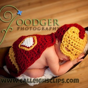 May include: A baby wearing a red and gold crocheted Iron Man costume, sleeping in a brown basket. The costume includes a cape and a mask. The image includes the text "GOODGER PHOTOGRAPHY" and "www.calleighsclips.com".