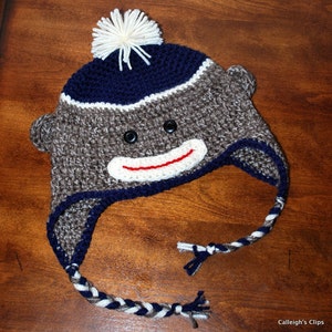May include: A brown and gray crocheted monkey hat with a white pom pom on top. The hat has a blue and white striped brim and a red smile.