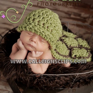 Puede incluir: Un bebé recién nacido con un gorro de tortuga de ganchillo verde y un atuendo a juego. El bebé duerme plácidamente en una cesta forrada de piel marrón. La imagen incluye el texto "GOODGER PHOTOGRAPHY" y "www.calleightsclips.com".