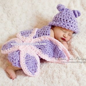 May include: A sleeping baby wearing a purple and pink crocheted butterfly costume with a matching hat. The costume has a large butterfly wing design with pink accents. The hat has two small ear-like flaps.