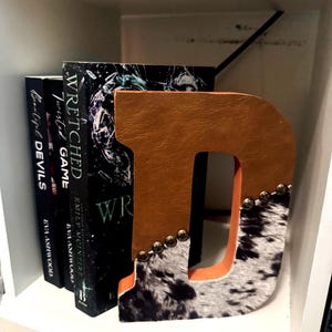 May include: A decorative letter "D" bookend with a brown leather front and a cowhide pattern on the bottom. The bookend is embellished with small gold-colored studs. Books with dark covers are visible behind the bookend.