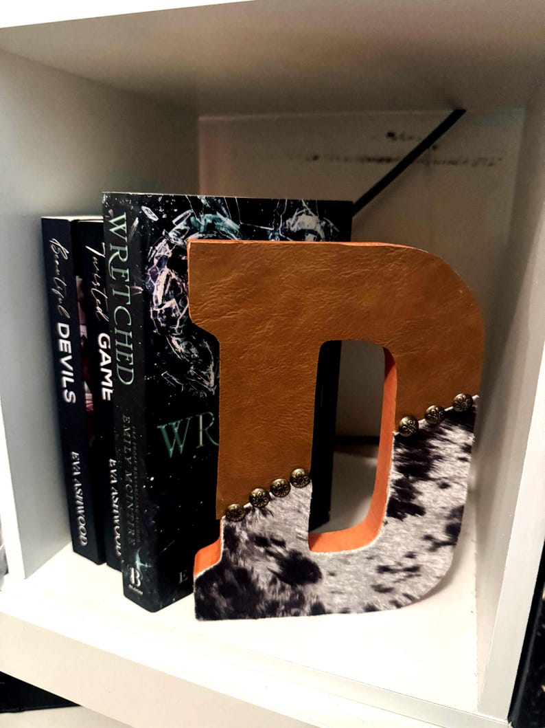 May include: A decorative letter "D" bookend with a brown leather front and a cowhide pattern on the bottom. The bookend is embellished with small gold-colored studs. Books with dark covers are visible behind the bookend.
