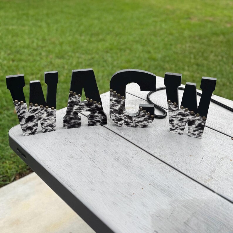 May include: Wooden letters spelling "WACW" with a black and cowhide pattern. The letters are on a white table with a green grassy background. The letters are decorated with small gold-colored studs.