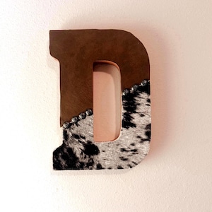 May include: A decorative letter "D" made of brown leather and cowhide. The top half is brown leather, while the bottom half features a black and white cowhide pattern. Silver studs line the edge where the materials meet.
