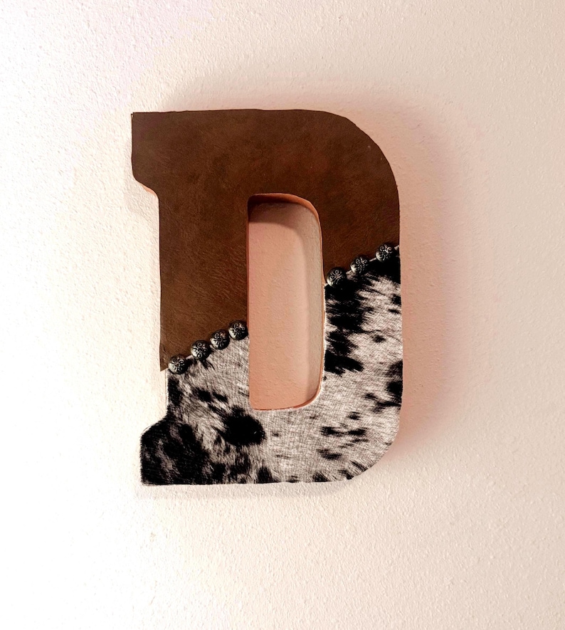 May include: A decorative letter "D" made of brown leather and cowhide. The top half is brown leather, while the bottom half features a black and white cowhide pattern. Silver studs line the edge where the materials meet.