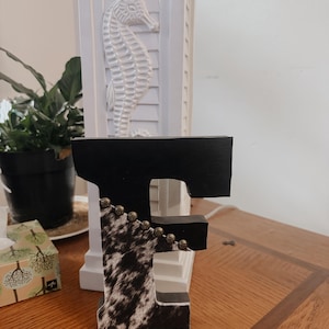 May include: A decorative wooden letter "F" with a black top and cowhide pattern on the front. The letter is adorned with small, round, gold-colored beads. The letter is displayed on a wooden surface.