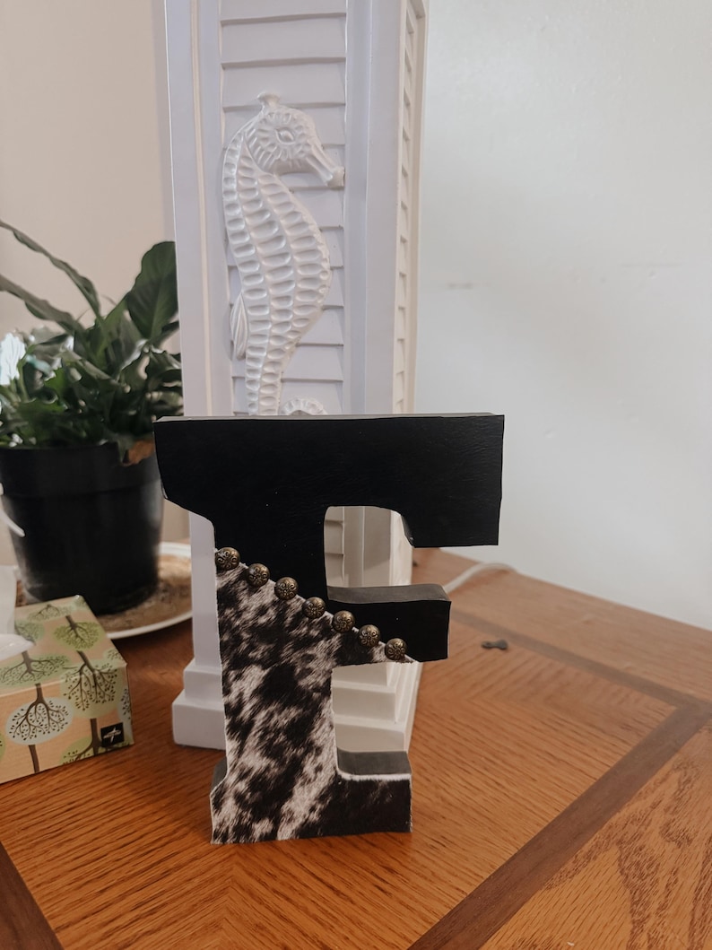 May include: A decorative wooden letter "F" with a black top and cowhide pattern on the front. The letter is adorned with small, round, gold-colored beads. The letter is displayed on a wooden surface.