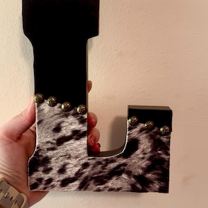 May include: A decorative wooden letter "L" with a black top and cowhide pattern on the front. The letter is adorned with small, gold-colored studs. The item is held against a neutral-colored wall.