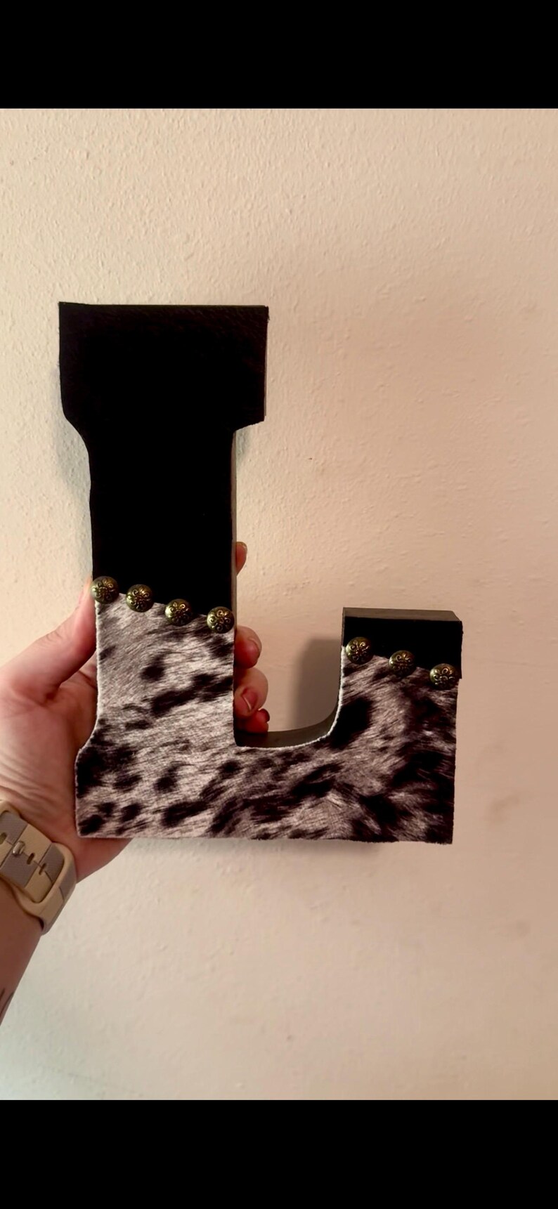 May include: A decorative wooden letter "L" with a black top and cowhide pattern on the front. The letter is adorned with small, gold-colored studs. The item is held against a neutral-colored wall.