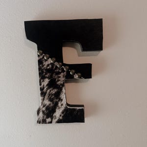 May include: A decorative wooden letter "E" with a cowhide pattern and black accents. The top and right side of the letter are black, while the left side features a black and white cowhide design. Small silver studs adorn the edge.
