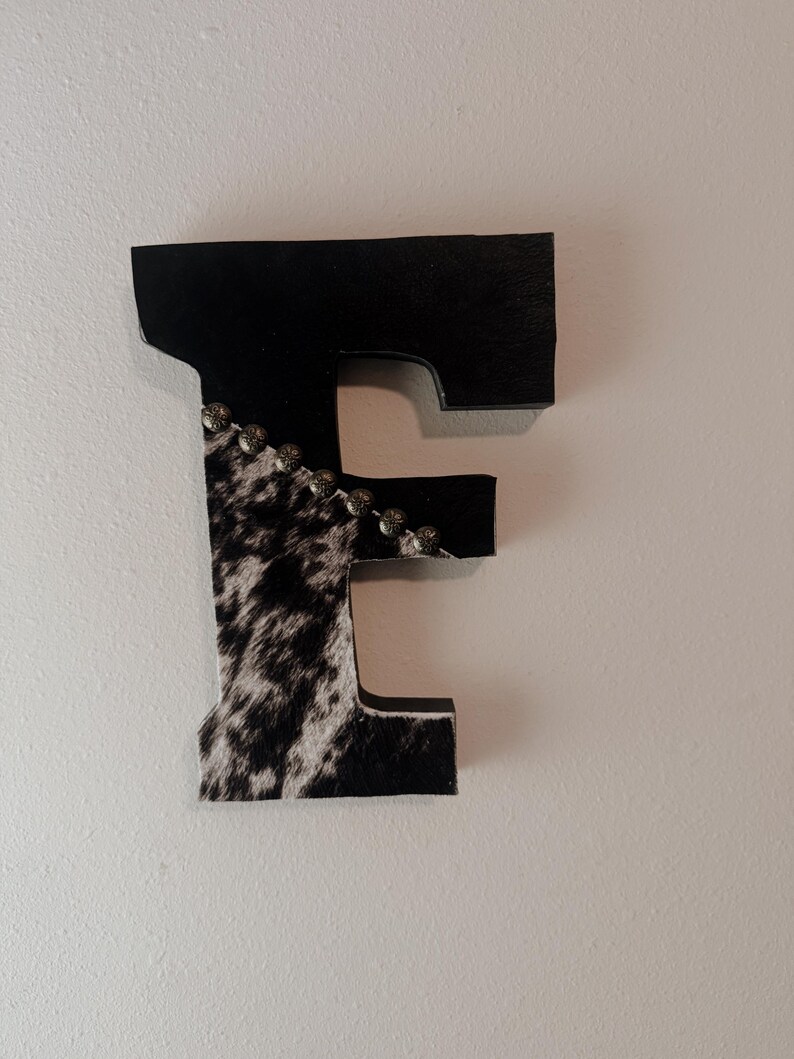 May include: A decorative wooden letter "E" with a cowhide pattern and black accents. The top and right side of the letter are black, while the left side features a black and white cowhide design. Small silver studs adorn the edge.
