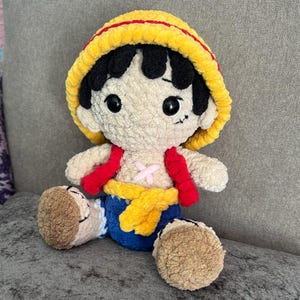 Puede incluir: Muñeco de peluche hecho a mano de un personaje con cabello negro, un sombrero amarillo con una raya roja y una cicatriz debajo del ojo izquierdo. El muñeco tiene un chaleco rojo, una faja amarilla y pantalones cortos azules. El muñeco está sentado sobre una superficie gris.