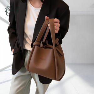 Peut inclure: Un sac à main en cuir marron avec une poignée supérieure et une fermeture à cordon. Le sac a une forme structurée et un petit logo en relief sur le devant. Le sac est tenu par une personne portant un blazer noir et un pantalon clair.