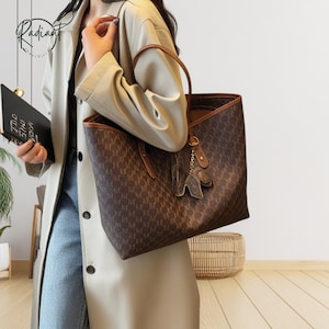 Könnte beinhalten: Braune Tragetasche mit sich wiederholendem Muster und braunen Ledergriffen. Die Tasche hat ein passendes braunes Innenfutter und einen dekorativen Anhänger. Die Tasche wird von einer Person getragen, die einen hellen Mantel und blaue Jeans trägt.