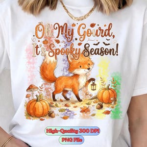 Peut inclure: Un t-shirt blanc avec un motif de renard tenant une lanterne, entouré de citrouilles, de champignons et de feuilles d'automne. Le texte indique "Oh My Gourd, It's Spooky Season!" et "High-Quality 300 DPI PNG File" en bas de l'image.