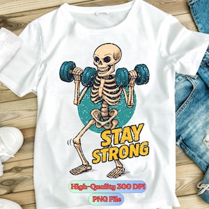 May include: White t-shirt featuring a cartoon skeleton lifting blue dumbbells. The skeleton is set against a teal circle, with the words "STAY STRONG" in yellow. The shirt has a distressed, vintage look.