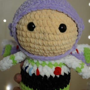 Puede incluir: Mu&ntilde;eca de ganchillo hecha a mano que se asemeja a un guardabosques espacial. La mu&ntilde;eca presenta un casco de color p&uacute;rpura claro, una cara de color beige con ojos negros y un traje espacial blanco y verde con detalles en negro y rojo. La mu&ntilde;eca es suave.