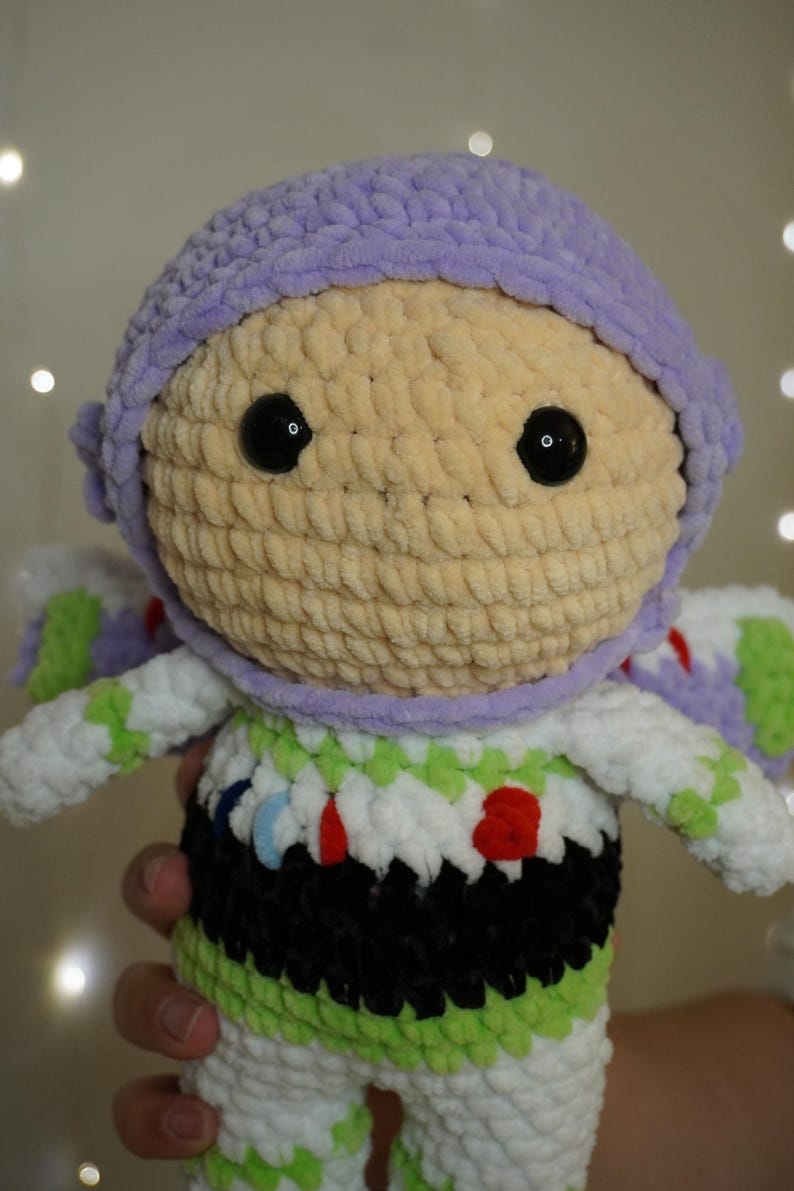 Puede incluir: Mu&ntilde;eca de ganchillo hecha a mano que se asemeja a un guardabosques espacial. La mu&ntilde;eca presenta un casco de color p&uacute;rpura claro, una cara de color beige con ojos negros y un traje espacial blanco y verde con detalles en negro y rojo. La mu&ntilde;eca es suave.