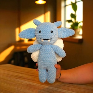 May include: A light blue crocheted plush toy in the shape of a friendly monster. The toy has large ears, white wings, and a smiling face with small fangs. The plush is held in a person's hand.