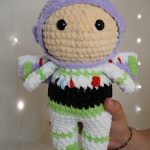 May include: A handmade crochet doll of a space ranger character. The doll features a light purple helmet, a beige face, and large black eyes. The outfit is white with green and black accents. The doll is approximately 10 inches tall.