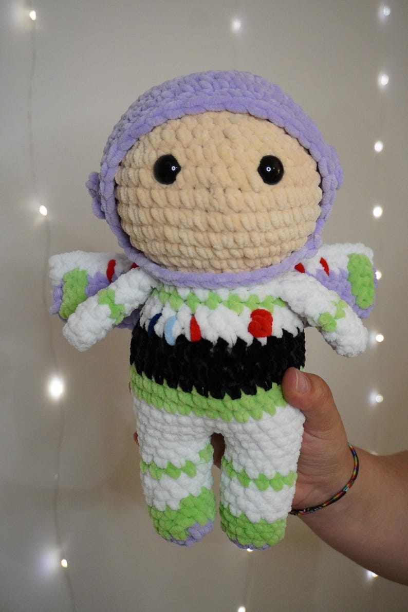 Puede incluir: Mu&ntilde;eca de ganchillo hecha a mano de un personaje de guardabosques espacial. La mu&ntilde;eca presenta un casco de color lila claro, una cara beige y ojos negros grandes. El atuendo es blanco con detalles en verde y negro. La mu&ntilde;eca mide aproximadamente 25 cm de alto.
