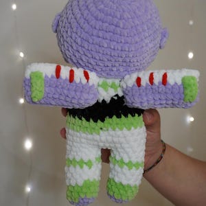 Puede incluir: Mu&ntilde;eco de ganchillo hecho a mano de un personaje de una popular pel&iacute;cula de animaci&oacute;n. El mu&ntilde;eco presenta una cabeza lavanda, pantalones a rayas blancas y verdes y brazos blancos y morados con detalles rojos. El mu&ntilde;eco es sostenido por una persona.