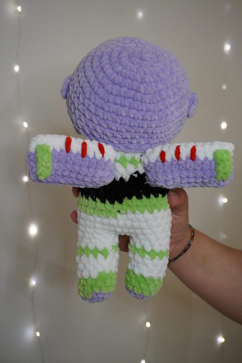 Puede incluir: Mu&ntilde;eco de ganchillo hecho a mano de un personaje de una popular pel&iacute;cula de animaci&oacute;n. El mu&ntilde;eco presenta una cabeza lavanda, pantalones a rayas blancas y verdes y brazos blancos y morados con detalles rojos. El mu&ntilde;eco es sostenido por una persona.