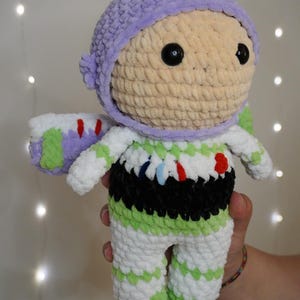 Puede incluir: Mu&ntilde;eca de ganchillo hecha a mano que se asemeja a Buzz Lightyear de Toy Story. La mu&ntilde;eca presenta un casco morado claro, una cara beige y un traje espacial blanco y verde con detalles en negro. La mu&ntilde;eca se sostiene frente a un fondo borroso.