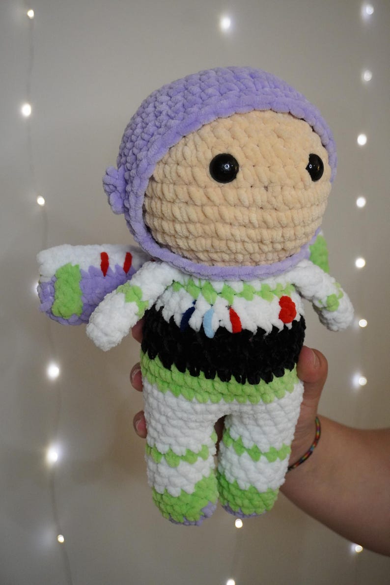 Puede incluir: Mu&ntilde;eca de ganchillo hecha a mano que se asemeja a Buzz Lightyear de Toy Story. La mu&ntilde;eca presenta un casco morado claro, una cara beige y un traje espacial blanco y verde con detalles en negro. La mu&ntilde;eca se sostiene frente a un fondo borroso.