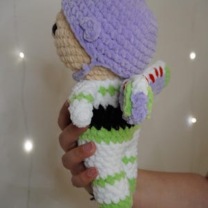 Puede incluir: Mu&ntilde;eca de ganchillo que se asemeja a un personaje de una popular pel&iacute;cula de animaci&oacute;n. La mu&ntilde;eca presenta un sombrero morado claro, una cabeza beige y un mono blanco y verde. Las alas de la mu&ntilde;eca son blancas, verdes y rojas. La mu&ntilde;eca est&aacute; sujeta en una mano.