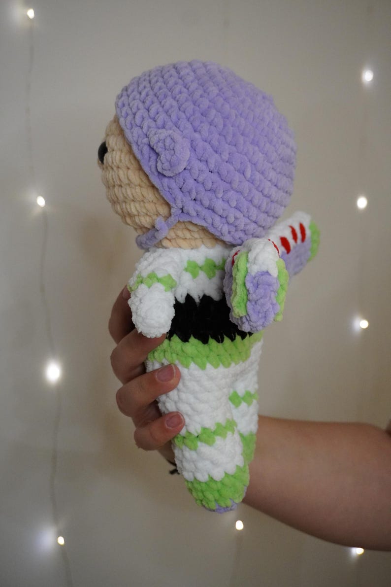 Puede incluir: Mu&ntilde;eca de ganchillo que se asemeja a un personaje de una popular pel&iacute;cula de animaci&oacute;n. La mu&ntilde;eca presenta un sombrero morado claro, una cabeza beige y un mono blanco y verde. Las alas de la mu&ntilde;eca son blancas, verdes y rojas. La mu&ntilde;eca est&aacute; sujeta en una mano.
