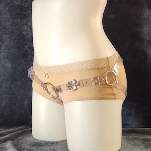 Peut inclure: Un ensemble de lingerie beige avec des bretelles en plastique transparent et des anneaux métalliques. L'ensemble comprend une culotte et un design de type harnais. La culotte a une bordure en dentelle sur le bord supérieur.