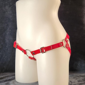 Könnte beinhalten: Roter Leder-Harness mit silberfarbenen Metallringen und Schnallen, präsentiert auf einem weißen Mannequin-Torso. Das Design umfasst einen zentralen Ring und verstellbare Riemen. Dunkler Hintergrund, Modeaccessoire.