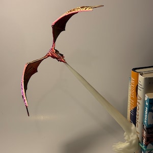 May include: A handcrafted, red and gold dragon sculpture with outstretched wings. The dragon appears to be breathing a white stream. The sculpture is positioned in front of a stack of books.