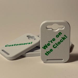 May include: White rectangular tags with rounded corners and a keyhole-shaped hole at the top. The text "Customers!" and "We're on the Clock!" are printed in green on the tags. The tags are stacked on a white surface.