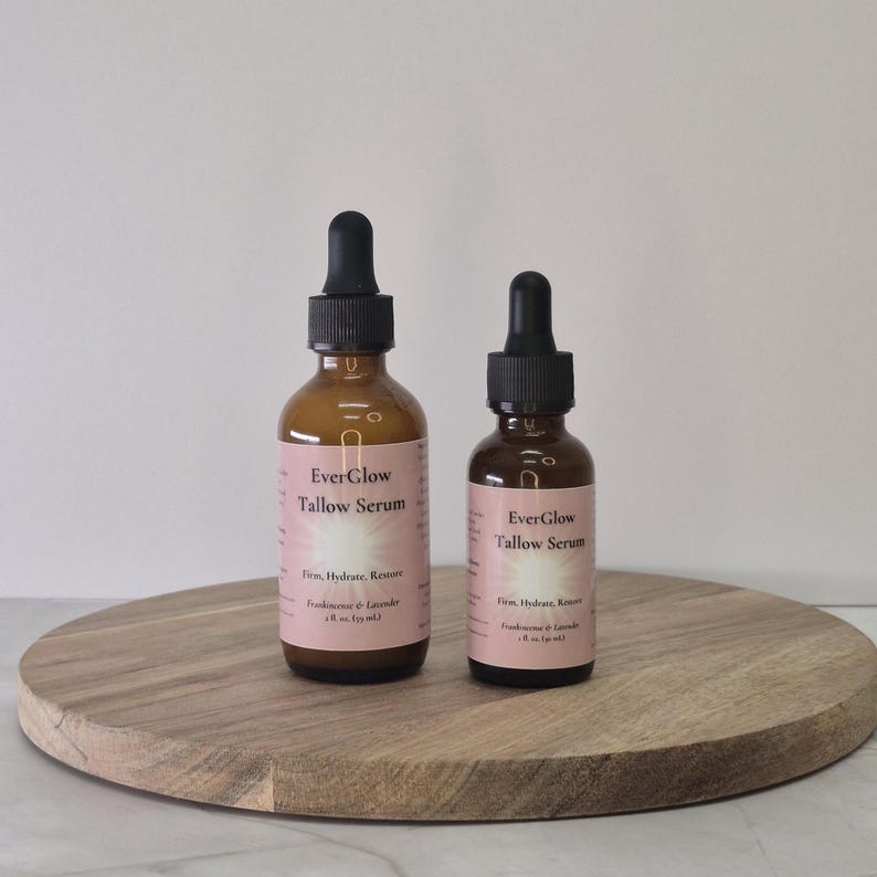 Op de afbeelding: Twee amberkleurige glazen flessen EverGlow Tallow Serum met zwarte druppelaars. De flessen hebben roze etiketten met witte tekst. De flessen staan op een ronde, lichtbruine houten tray.